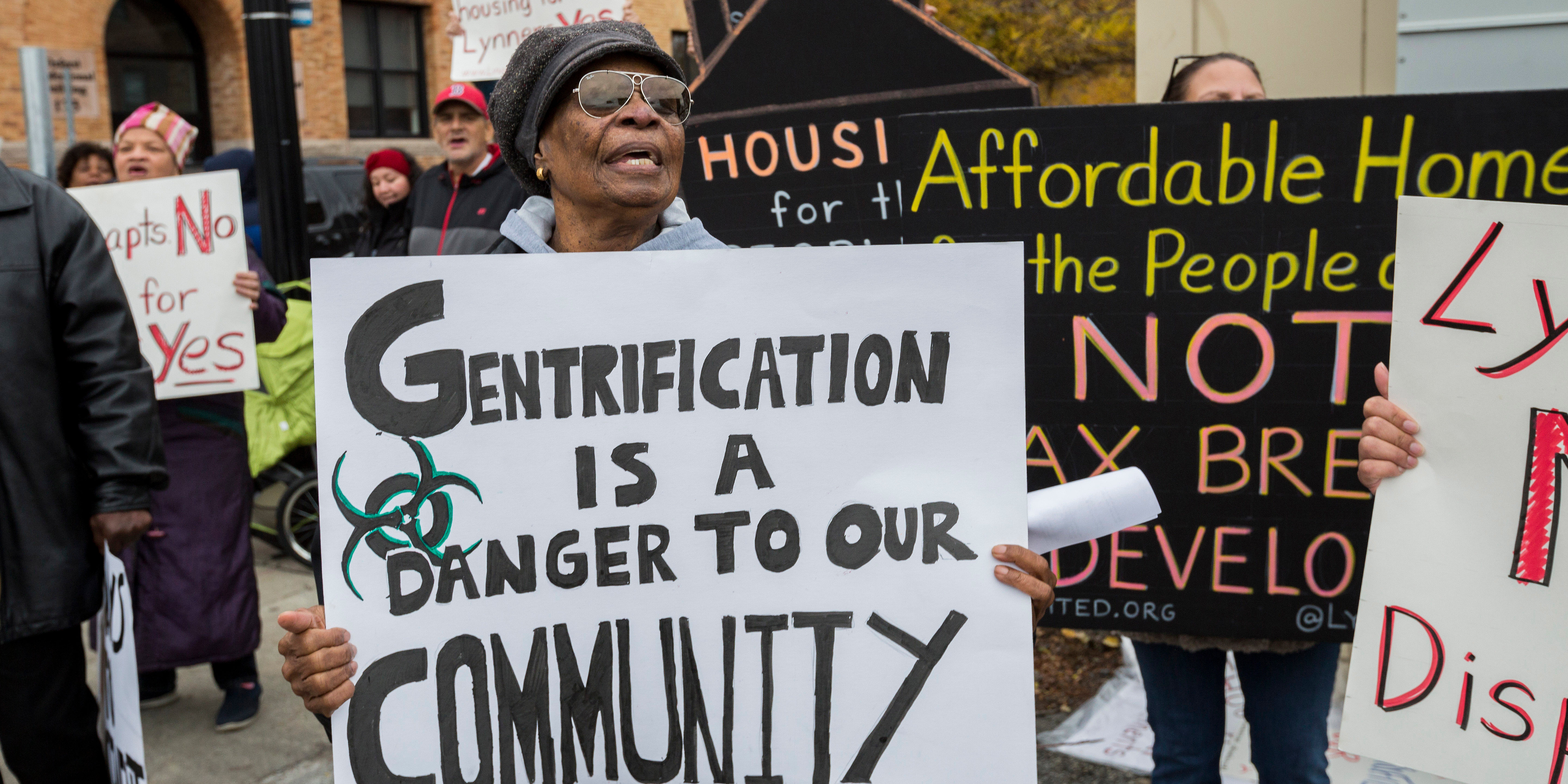Crowd with protest signs. Woman in foreground holds sign with a biohazard symbol and text reading Gentrification is a danger to our community.
